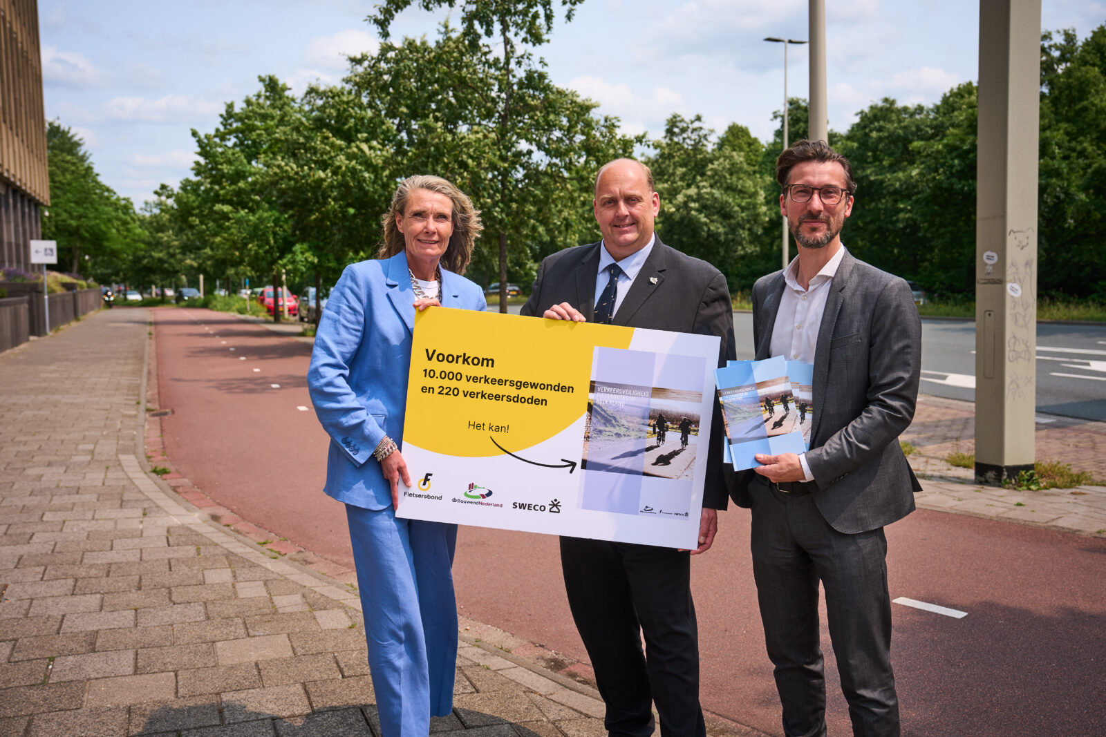 Esther van Garderen (Fietsersbond) en Hessel Heins-Wunderle (Bouwend Nederland) overhandigen het rapport over veiligheid fietsroutes in Nederland aan Frederik Zevenbergen (VVD), gedeputeerde Provincie Zuid-Holland. Op de achtergrond een fietspad.